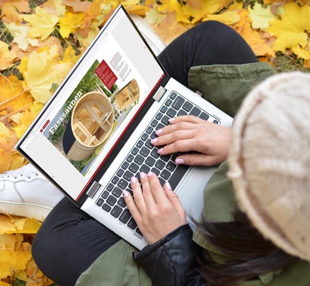 Frau sitzt im Schneidersitz in heruntergefallenem Herbstlaub mit Laptop auf den Beinen. Auf dem Bildschirm ist eine WEKA Fasssauna zu sehen.